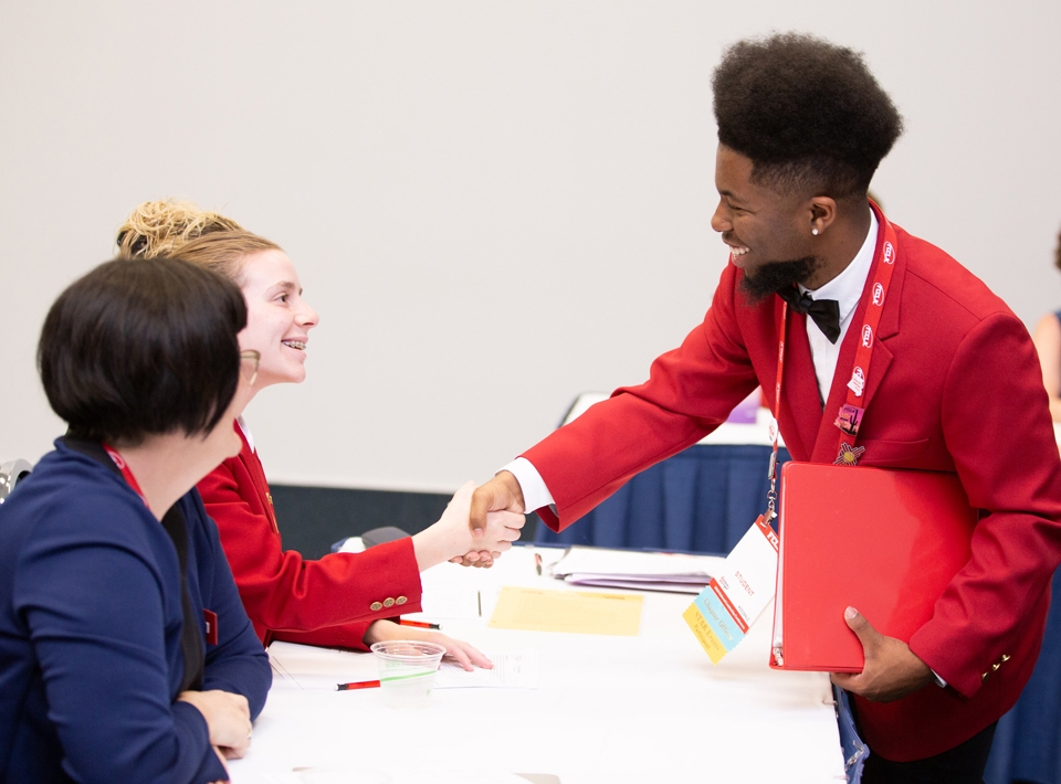 Image from the FCCLA homepage: a student in a red jacket shakes hands with 2 students sitting at a table.